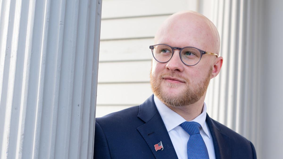 一个有胡子的, bald man with eyeglasses, wearing a suit with an American flag lapel pin stands near a Doric column.